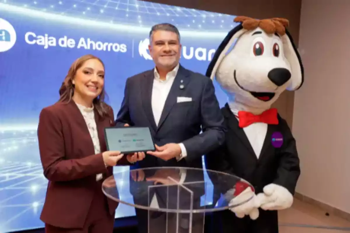 Dos personas posan sonrientes con una tableta en las manos junto a una botarga de perro con moño rojo, frente a un fondo azul con el logo de Caja de Ahorros.