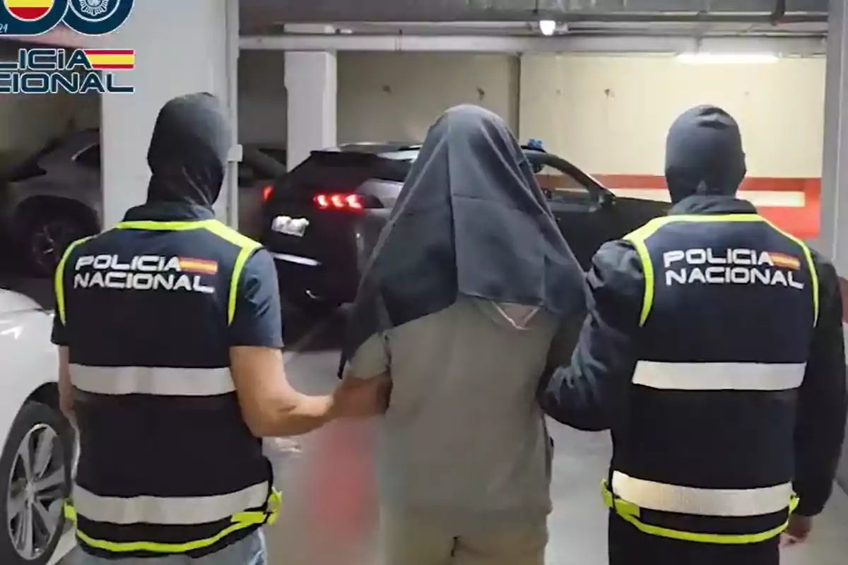 Dos agentes de la Policía Nacional escoltan a una persona detenida con la cabeza cubierta en un estacionamiento subterráneo.