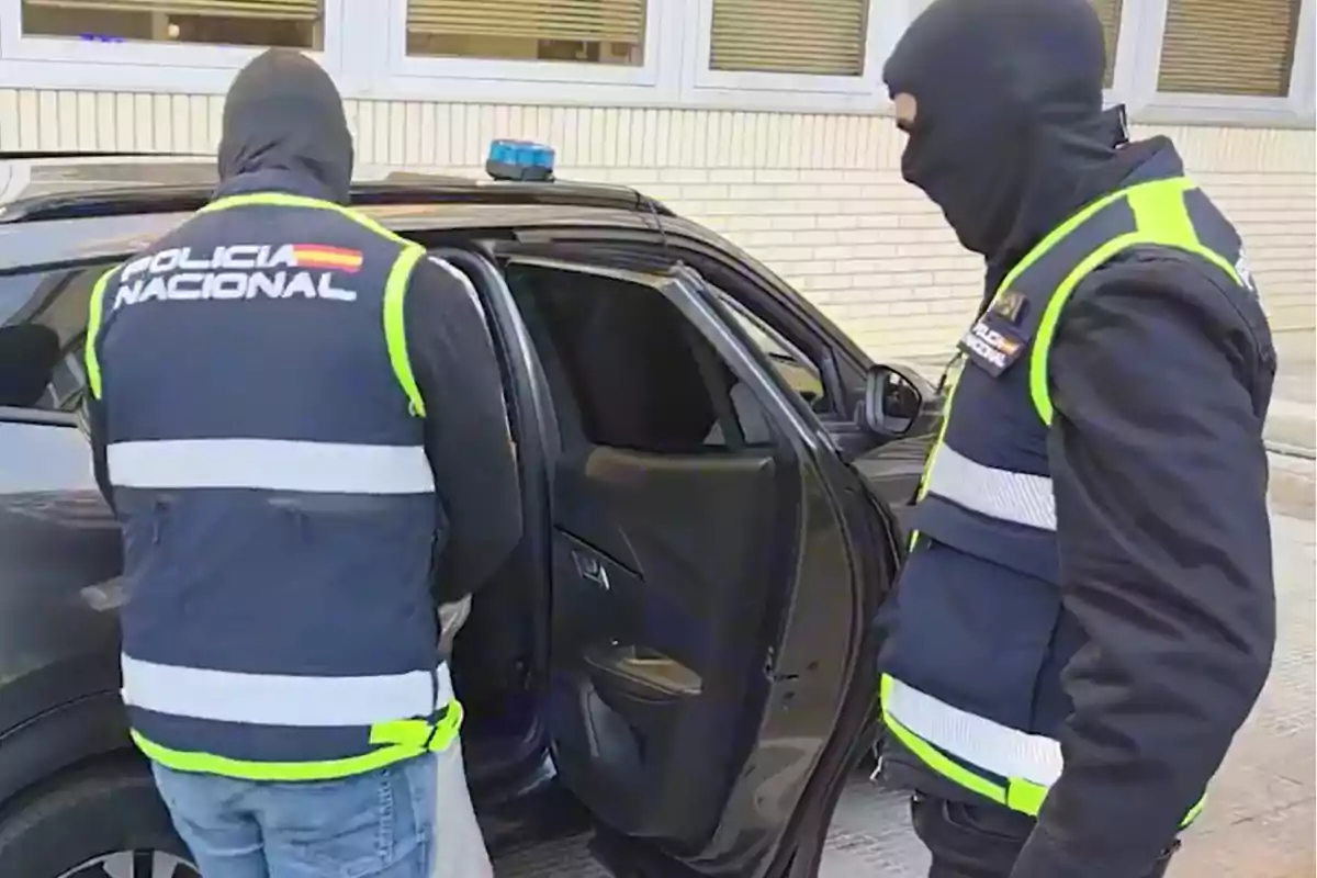 Dos agentes de la policía nacional con chalecos y pasamontañas junto a un coche patrulla con la puerta trasera abierta