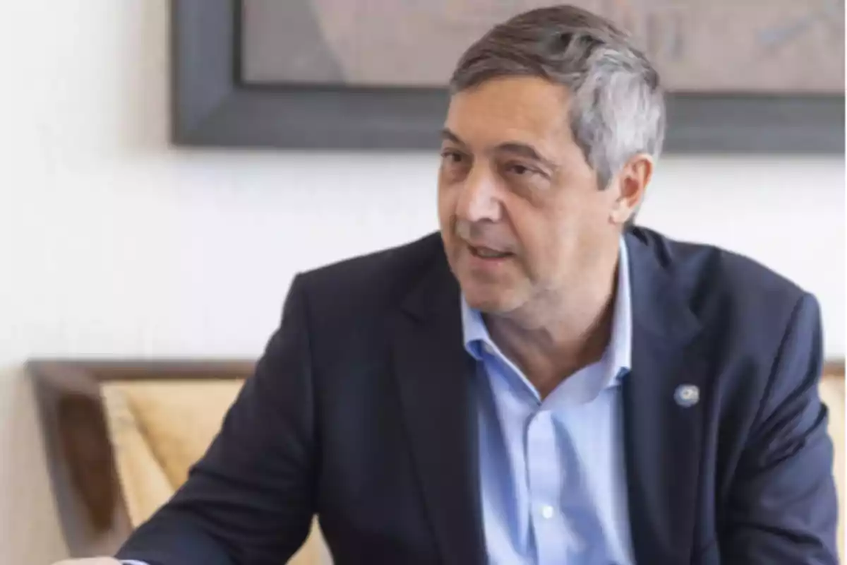 Hombre de cabello canoso con saco azul sentado en un sillón beige