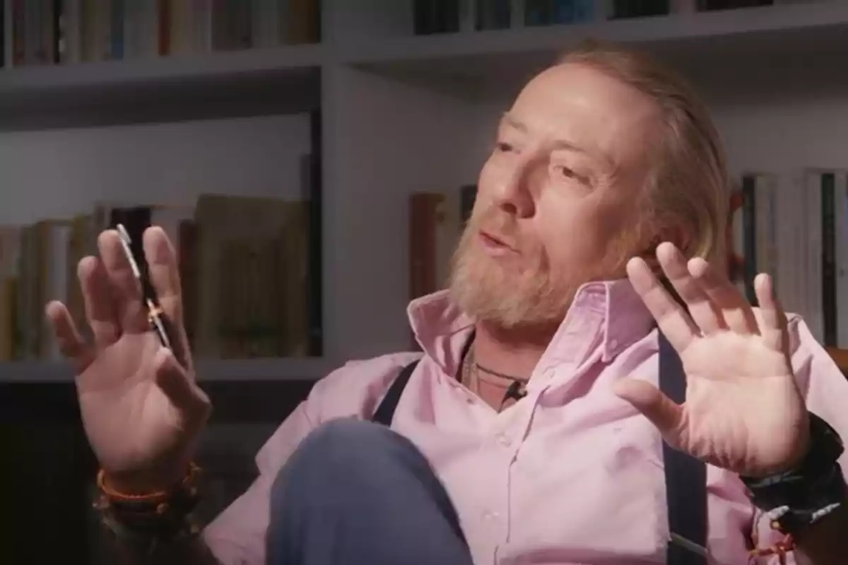 Hombre de barba rubia y camisa rosa gesticulando con las manos mientras sostiene una pluma, sentado frente a una estantería llena de libros