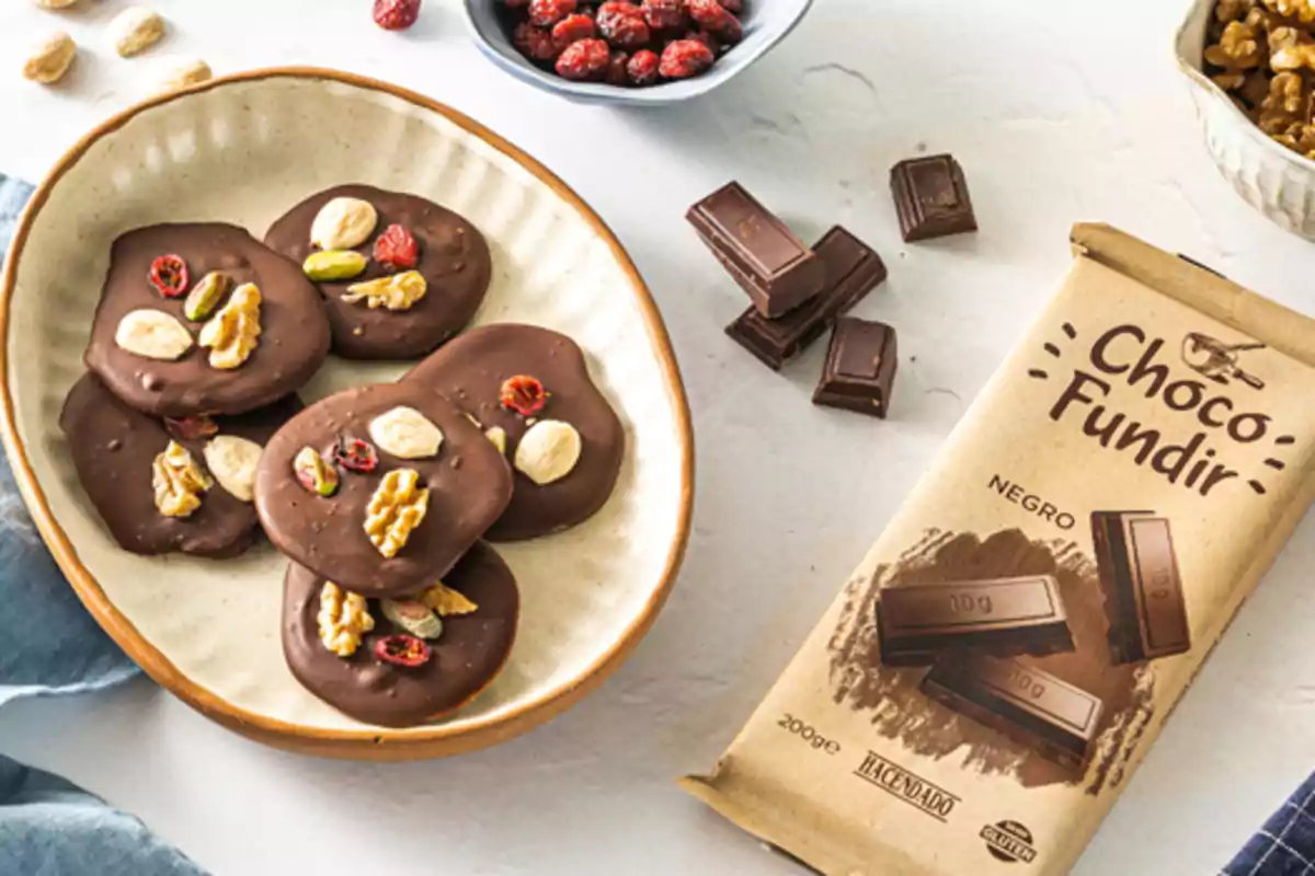 Tableta de chocolate negro junto a trozos de chocolate y un plato con discos de chocolate decorados con nueces, almendras, pistaches y arándanos secos