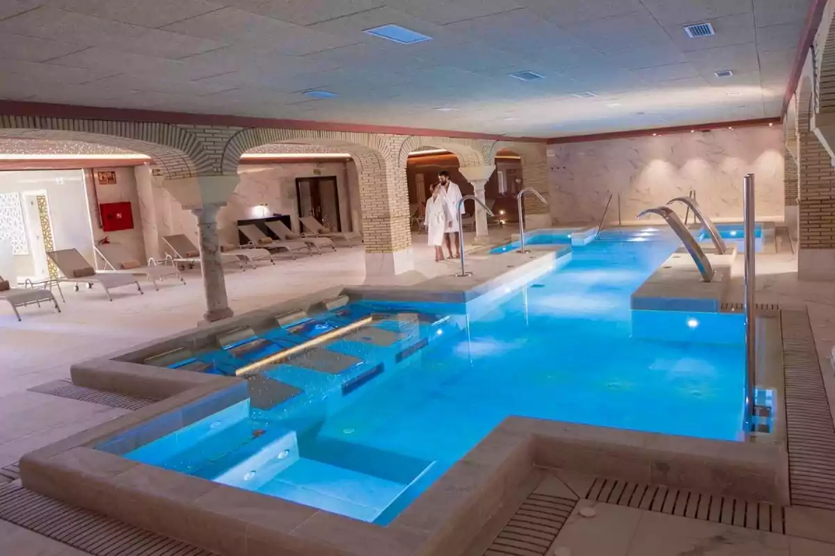 Piscina interior de un spa con agua azul, camastros alrededor y dos personas con batas blancas de baño de pie junto a la piscina