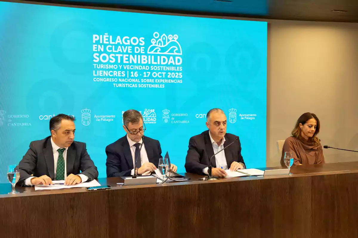 Cuatro personas sentadas en una mesa durante una conferencia sobre sostenibilidad turística en Piélagos con un fondo azul que muestra información del evento