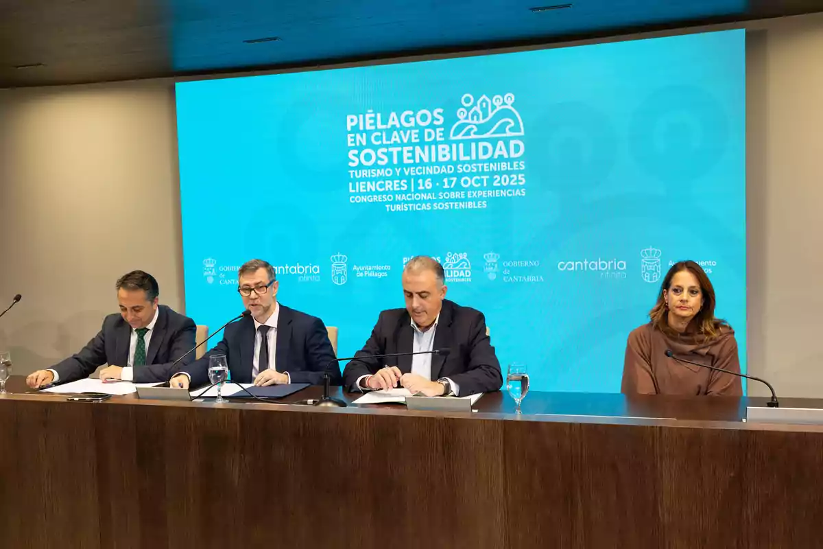 Cuatro personas sentadas en una mesa durante una conferencia sobre sostenibilidad turística con una pantalla azul de fondo que muestra información del evento en Piélagos