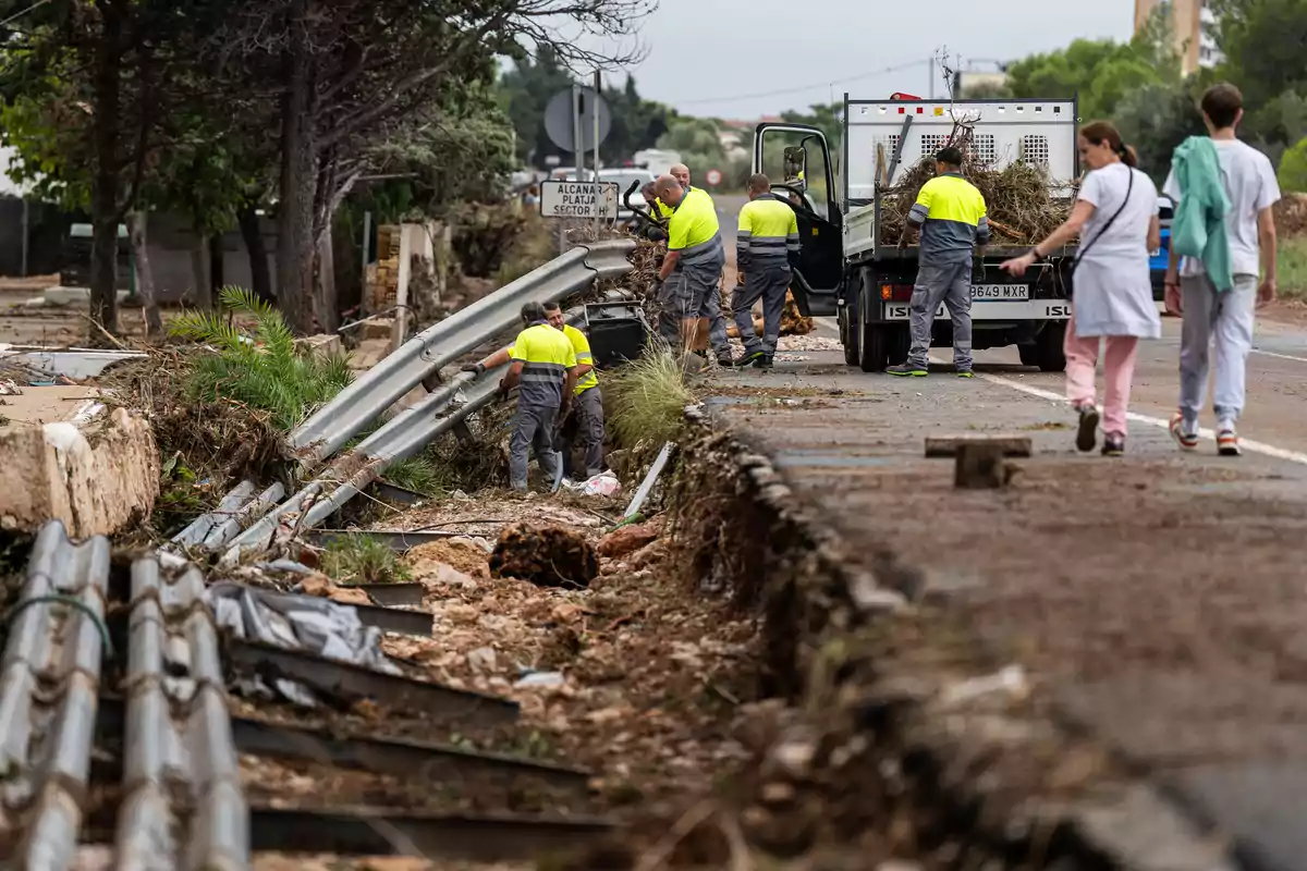Trabajadores con chalecos reflectantes reparan daños en una carretera mientras varias personas caminan cerca y se observa un camión de trabajo estacionado Trabajadores con chalecos reflectantes reparan daños en una carretera mientras varias personas caminan cerca y se observa un camión de trabajo estacionado