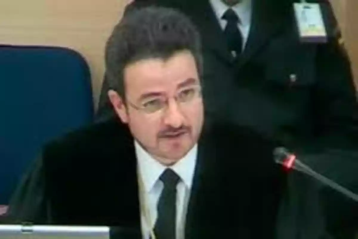 Hombre con gafas y bigote hablando frente a un micrófono en una sala formal