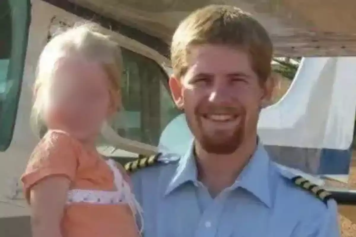 Un hombre con uniforme de piloto sonríe mientras sostiene a una niña con el rostro difuminado frente a una avioneta Un hombre con uniforme de piloto sonríe mientras sostiene a una niña con el rostro difuminado frente a una avioneta