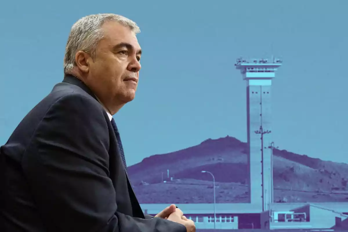 Hombre de cabello canoso con traje oscuro sentado en primer plano con una torre de control y una montaña al fondo