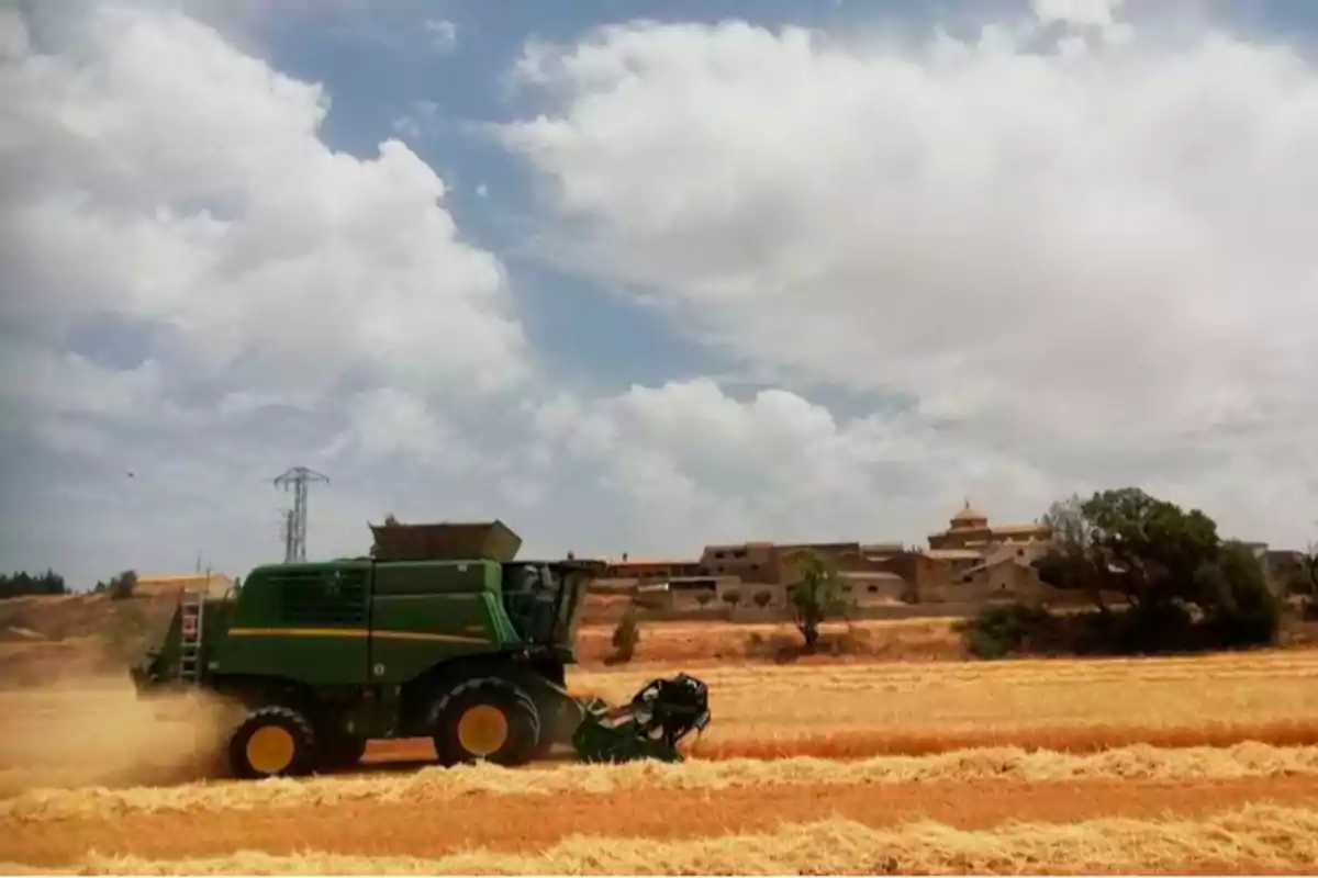 Cosechadora verde trabajando en un campo de trigo bajo un cielo parcialmente nublado con un pueblo al fondo