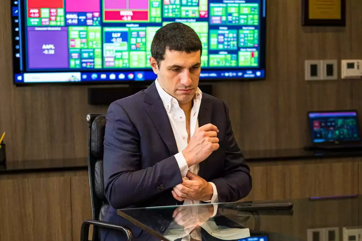 Hombre de cabello corto y oscuro con saco azul marino y camisa blanca sentado en una oficina moderna frente a una mesa de cristal con pantallas de datos financieros al fondo