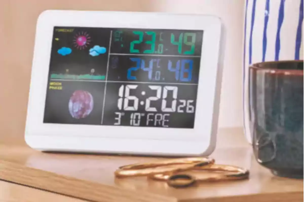 Reloj digital multifunción con pantalla a color que muestra la hora, temperatura, humedad, pronóstico del clima y fase lunar, colocado sobre una mesa junto a una taza y unas llaves doradas. Reloj digital multifunción con pantalla a color que muestra la hora, temperatura, humedad, pronóstico del clima y fase lunar, colocado sobre una mesa junto a una taza y unas llaves doradas.