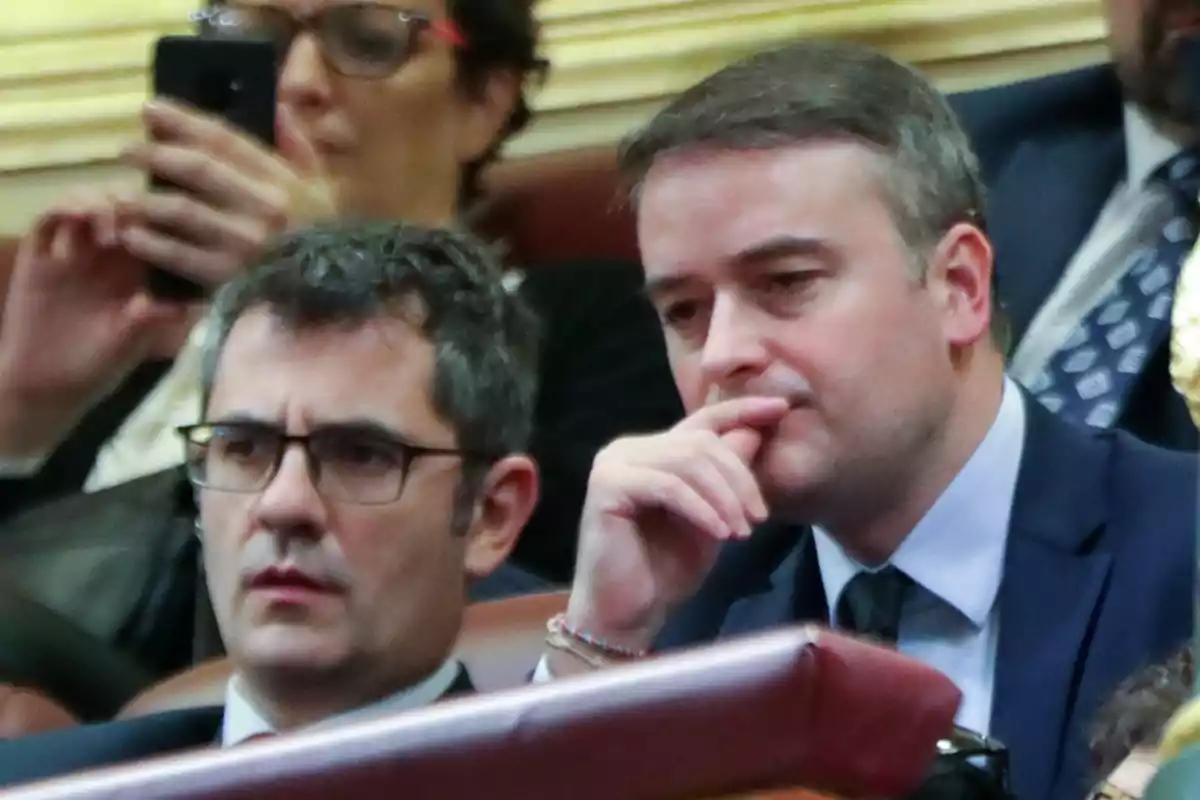 Dos hombres con traje y corbata sentados en lo que parece ser un recinto parlamentario, uno de ellos lleva gafas y ambos tienen expresión seria y pensativa