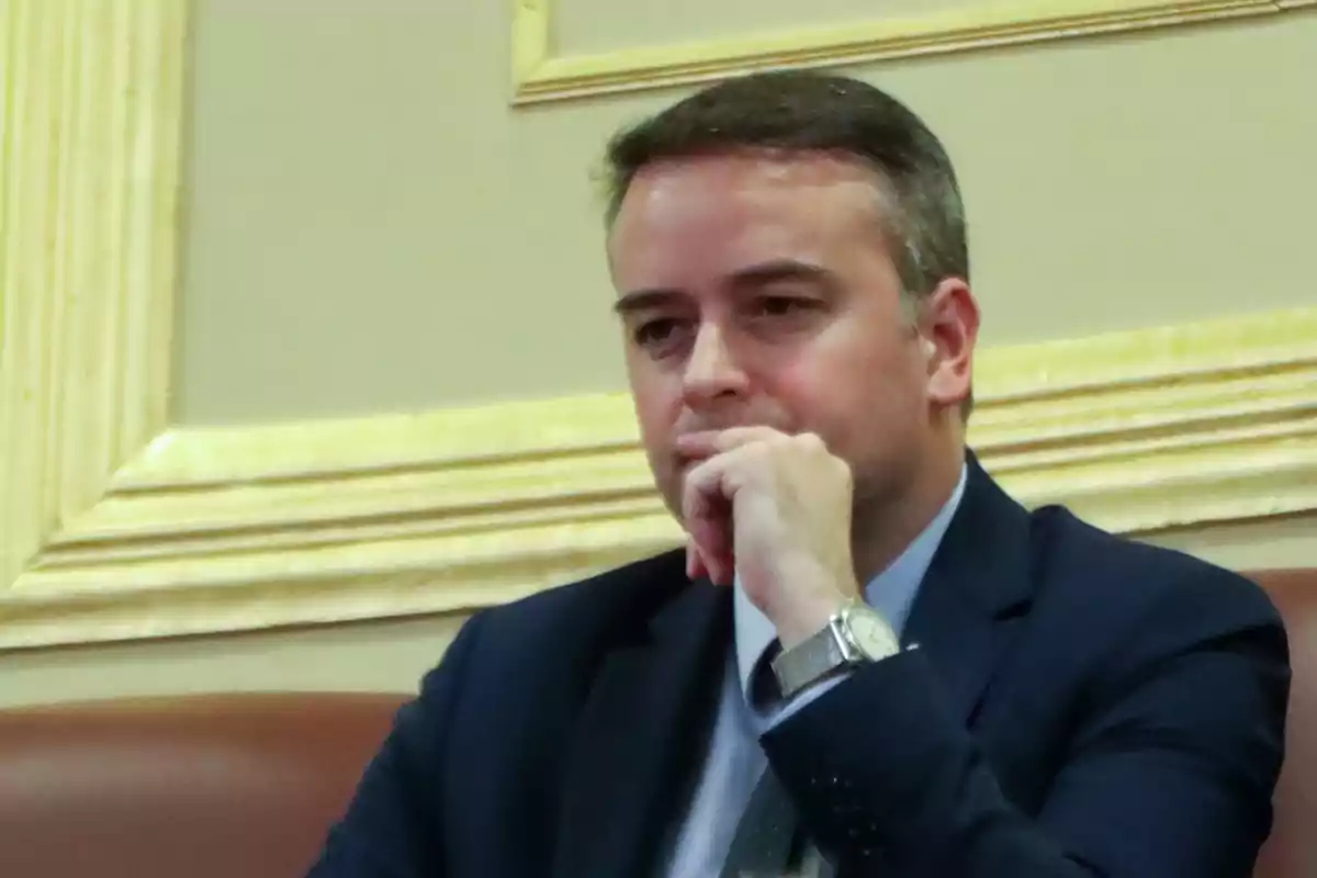 Hombre de traje oscuro sentado con expresión pensativa y la mano apoyada en el mentón Hombre de traje oscuro sentado con expresión pensativa y la mano apoyada en el mentón