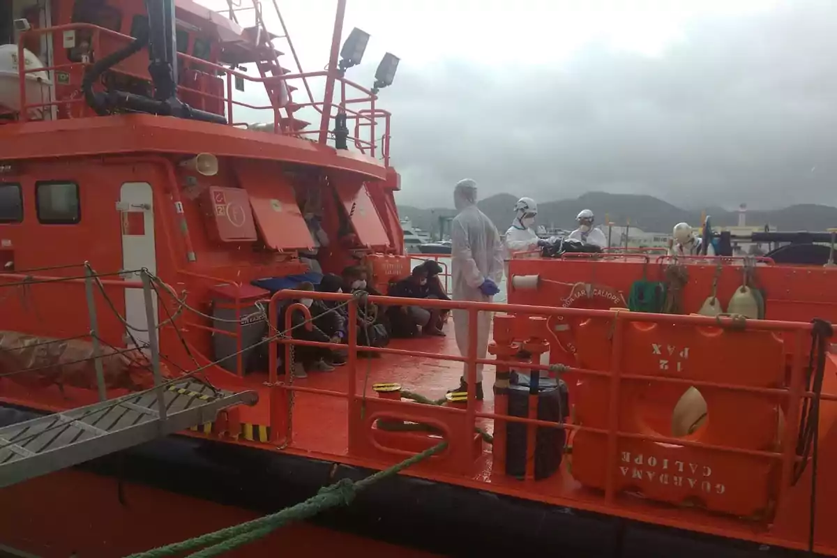 Personas con trajes de protección y mascarillas están en la cubierta de un barco de rescate naranja junto a varias personas sentadas, con un fondo de cielo nublado y montañas. Personas con trajes de protección y mascarillas están en la cubierta de un barco de rescate naranja junto a varias personas sentadas, con un fondo de cielo nublado y montañas.