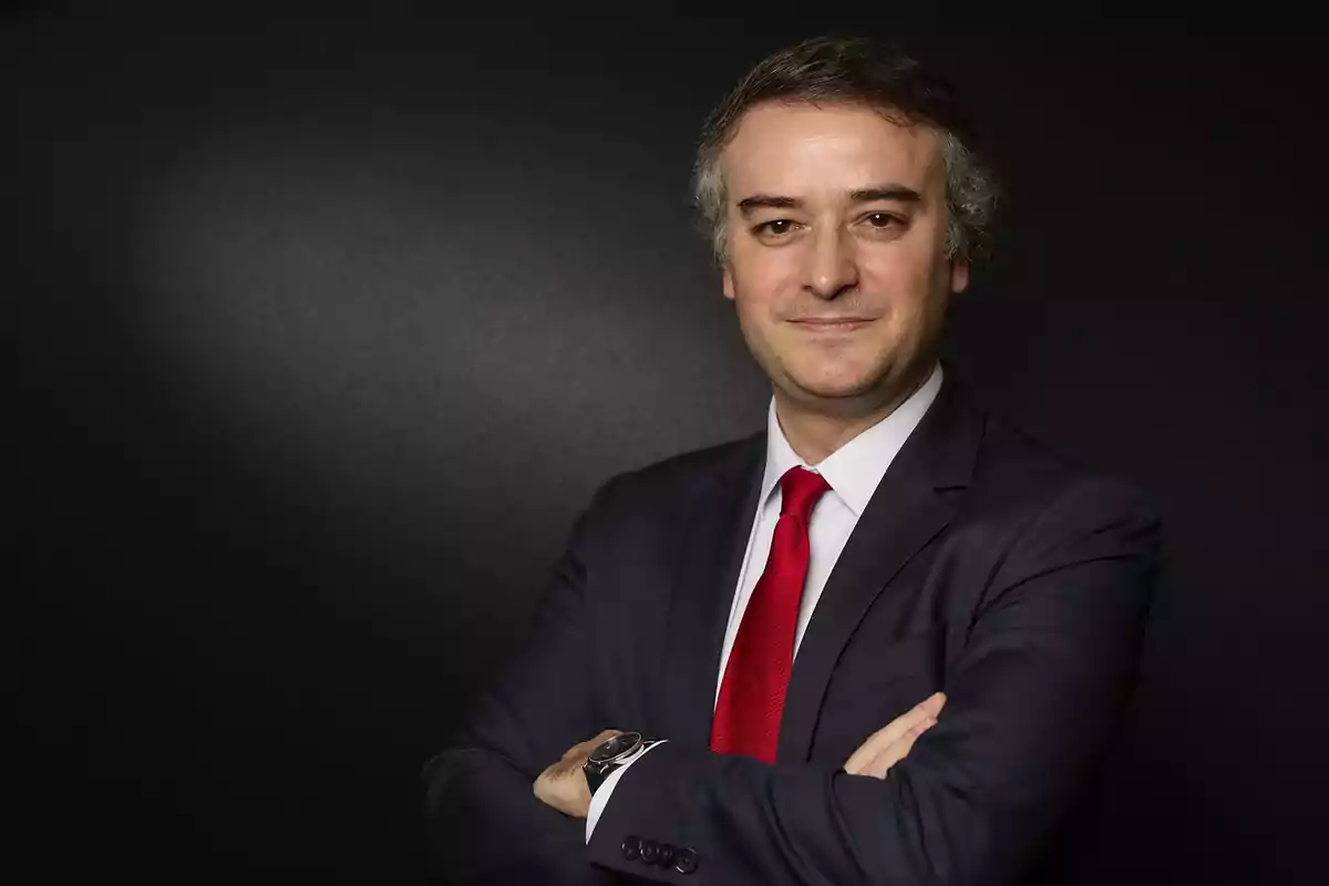 Hombre de cabello canoso con traje oscuro y corbata roja posando con los brazos cruzados frente a un fondo negro