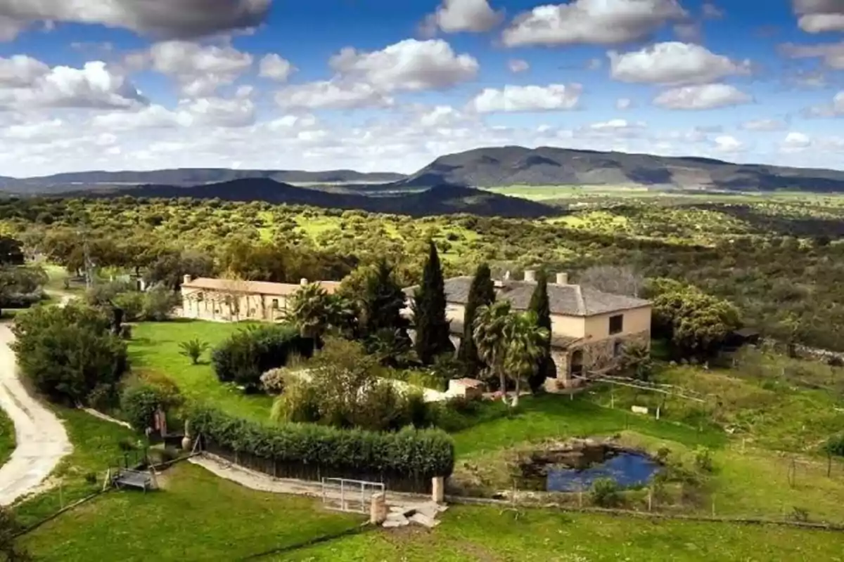 Vista aérea de una casa de campo rodeada de vegetación y colinas bajo un cielo azul con nubes dispersas