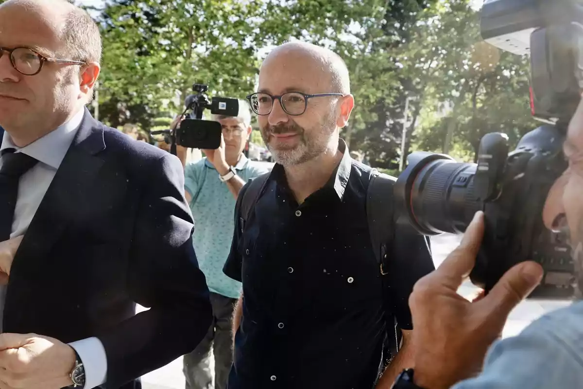 Un hombre con gafas y barba camina al aire libre mientras es fotografiado y grabado por periodistas