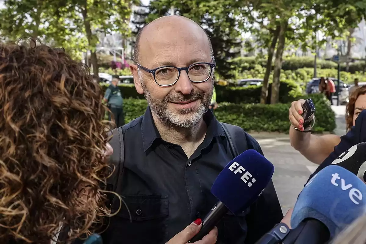 Hombre con gafas y barba siendo entrevistado por varios medios de comunicación en un entorno al aire libre con árboles y personas al fondo