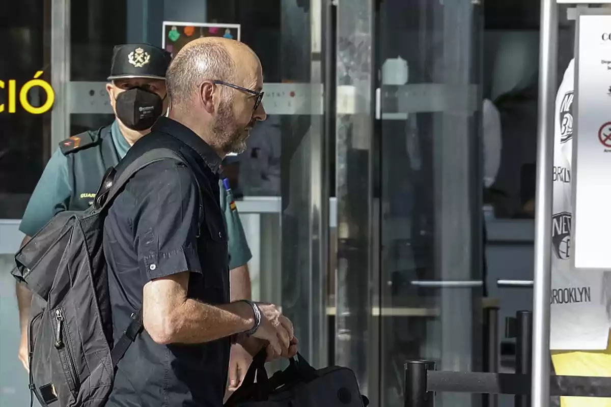 Un hombre con mochila y gafas camina frente a un guardia civil en la entrada de un edificio moderno