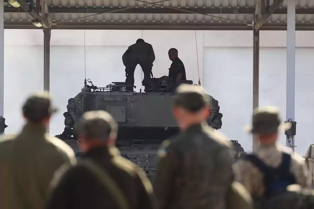 Varios soldados observan mientras dos personas están sobre un vehículo militar blindado bajo un techo metálico.