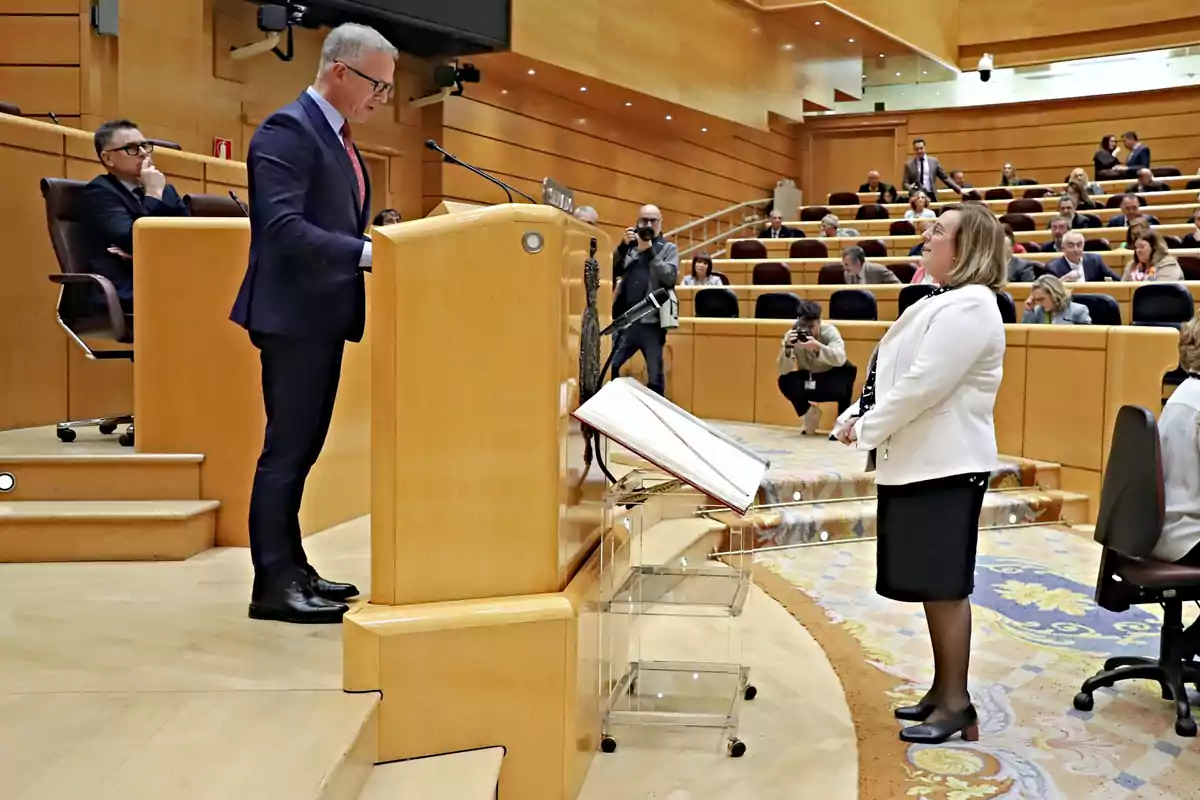 Un hombre de traje habla desde un atril en un recinto parlamentario mientras una mujer de pie lo escucha y varias personas observan y fotografían la escena desde sus asientos