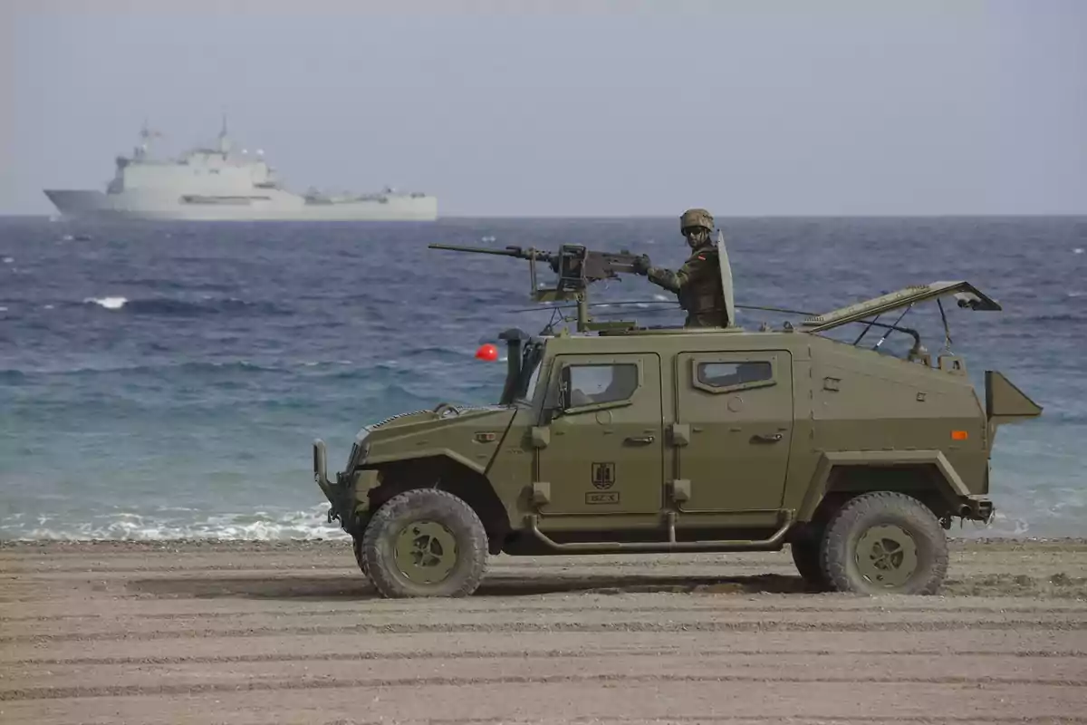 Vehículo militar blindado con un soldado armado en la playa y un barco de guerra en el mar al fondo