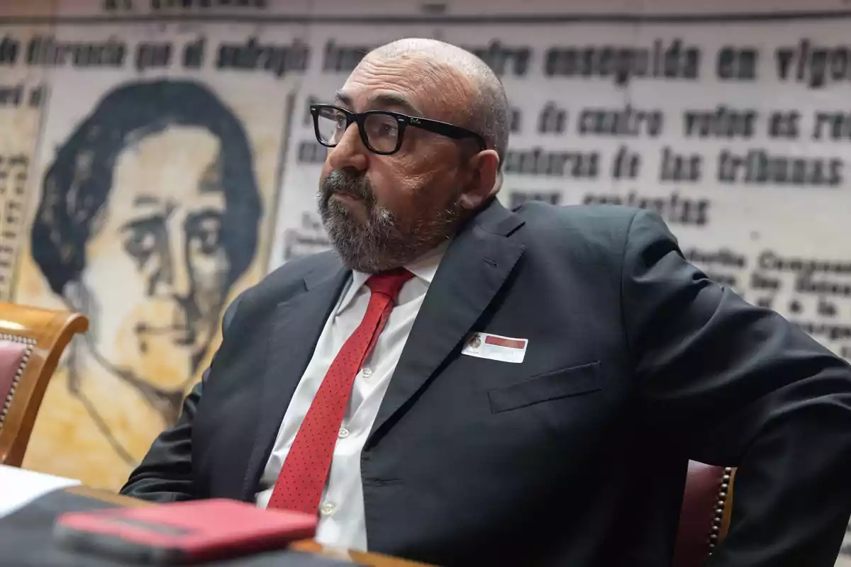 Hombre con barba y gafas vestido con traje oscuro y corbata roja sentado en una sala con fondo de mural y texto antiguo
