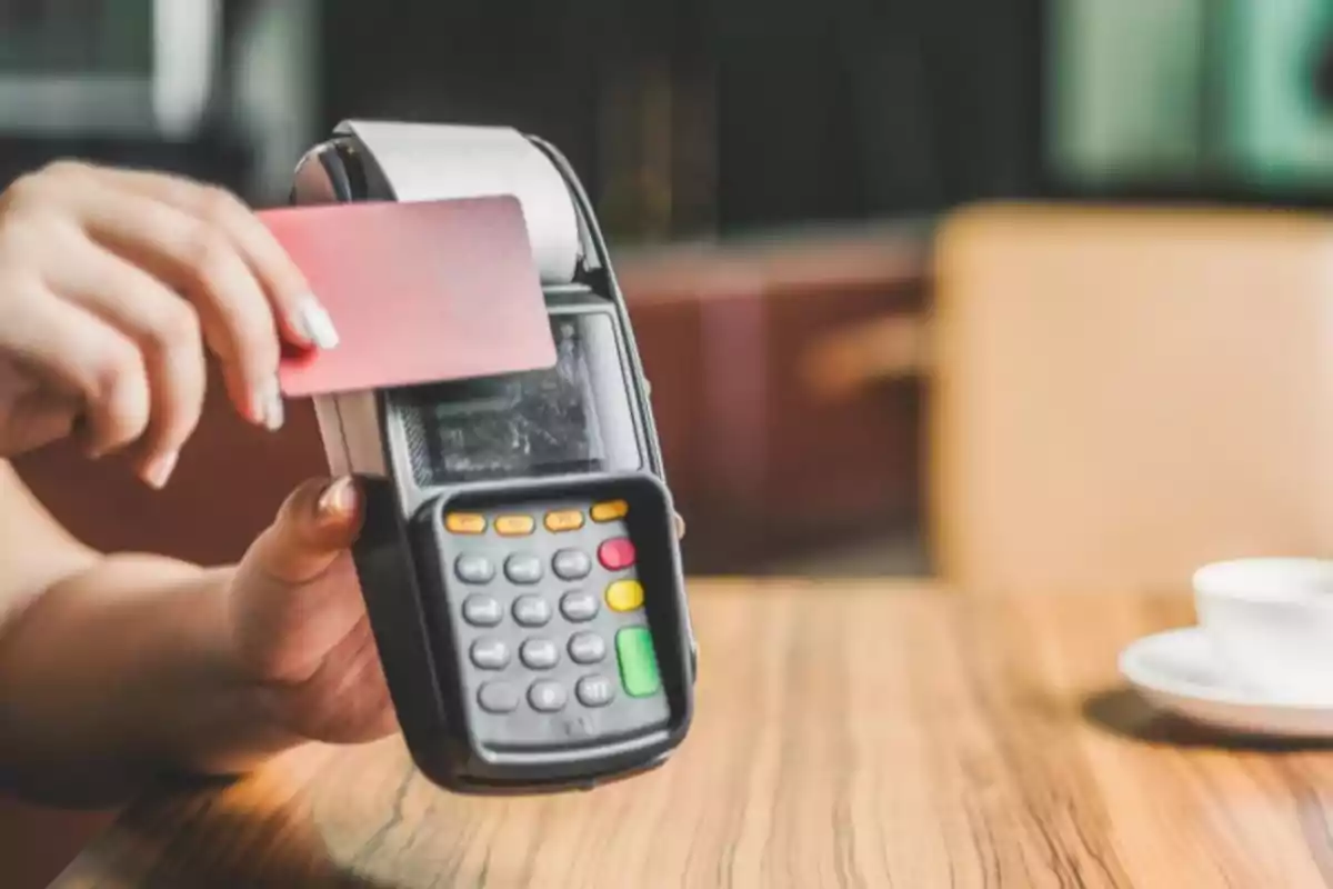 Persona deslizando una tarjeta bancaria en una terminal de pago sobre una mesa de madera Persona deslizando una tarjeta bancaria en una terminal de pago sobre una mesa de madera