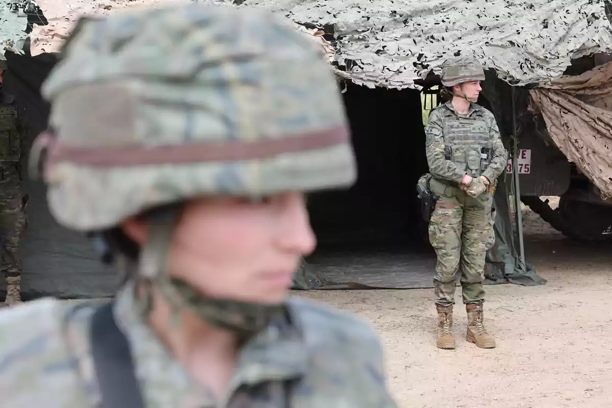 Dos mujeres militares con uniforme de camuflaje y casco, una en primer plano desenfocada y otra de pie al fondo frente a una tienda de campaña Dos mujeres militares con uniforme de camuflaje y casco, una en primer plano desenfocada y otra de pie al fondo frente a una tienda de campaña