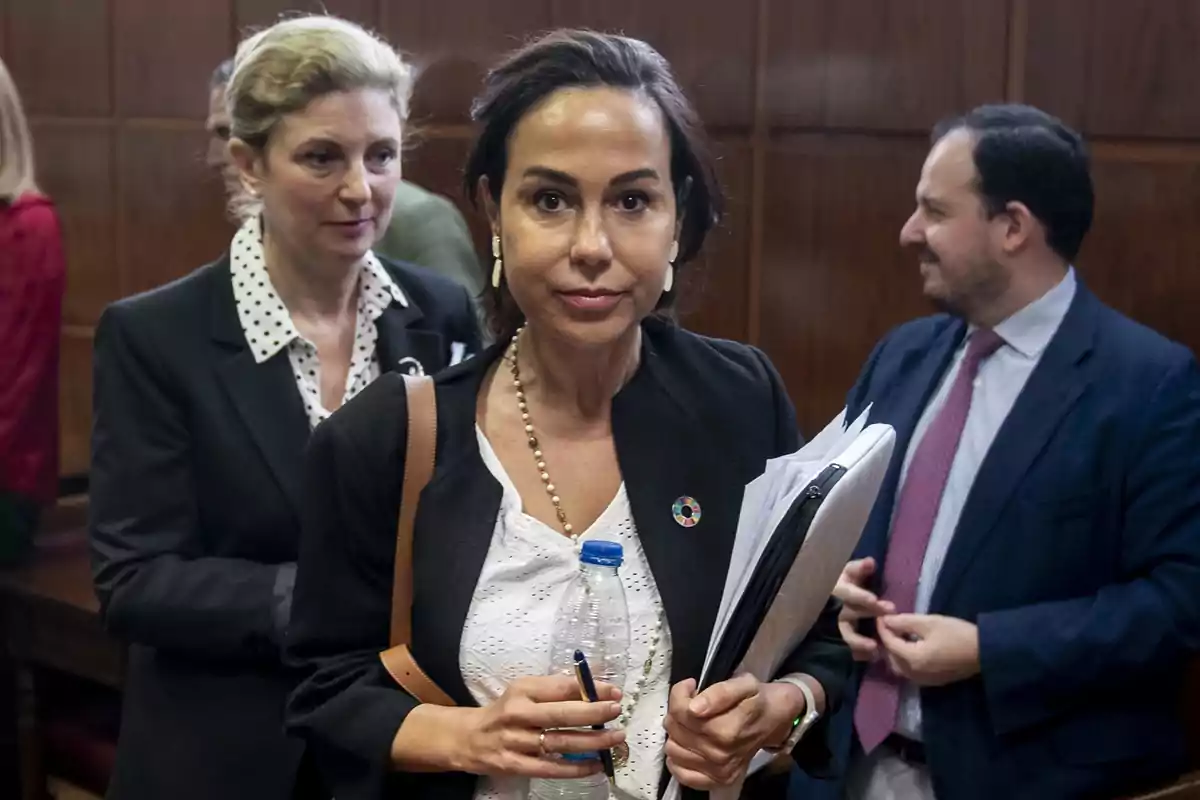 Mujer de cabello oscuro con chaqueta negra y blusa blanca sosteniendo una botella de agua y documentos, acompañada de otras personas en un entorno formal