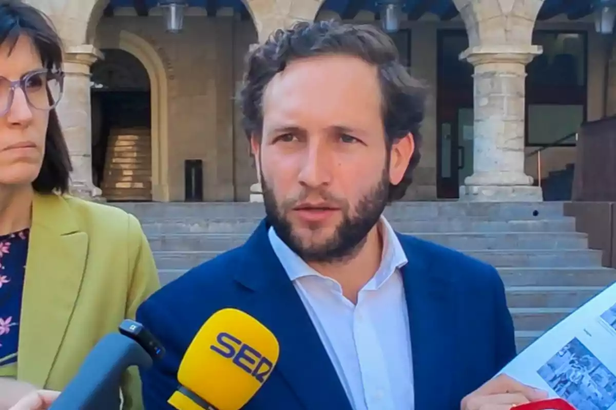 Un hombre con barba y traje azul habla frente a unos micrófonos mientras sostiene unos papeles, acompañado por una mujer con gafas y chaqueta amarilla, en unas escaleras de piedra con arcos al fondo.
