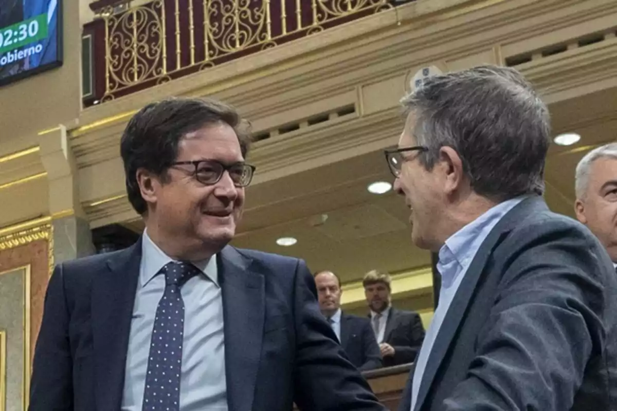 Dos hombres con gafas conversan y sonríen en un recinto parlamentario elegante con barandales dorados y otras personas al fondo Dos hombres con gafas conversan y sonríen en un recinto parlamentario elegante con barandales dorados y otras personas al fondo