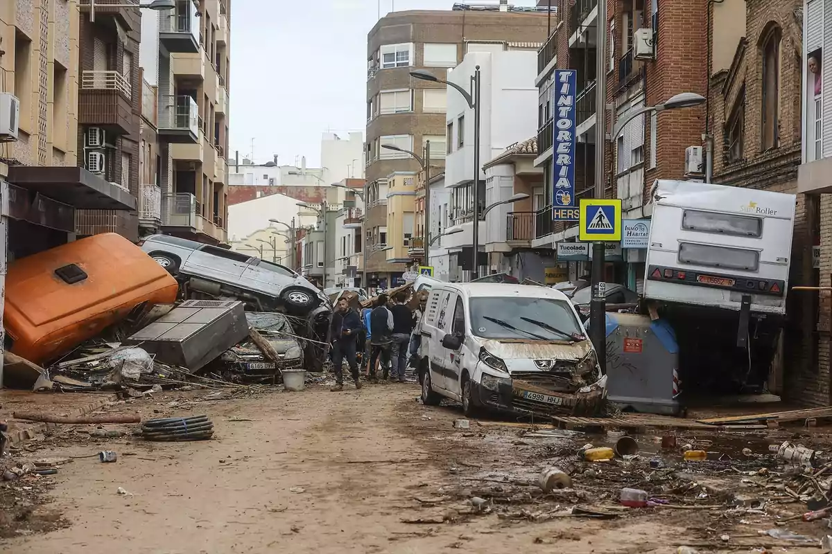 Calle urbana con varios vehículos dañados y apilados, escombros y basura tras una inundación, personas observando los daños entre edificios