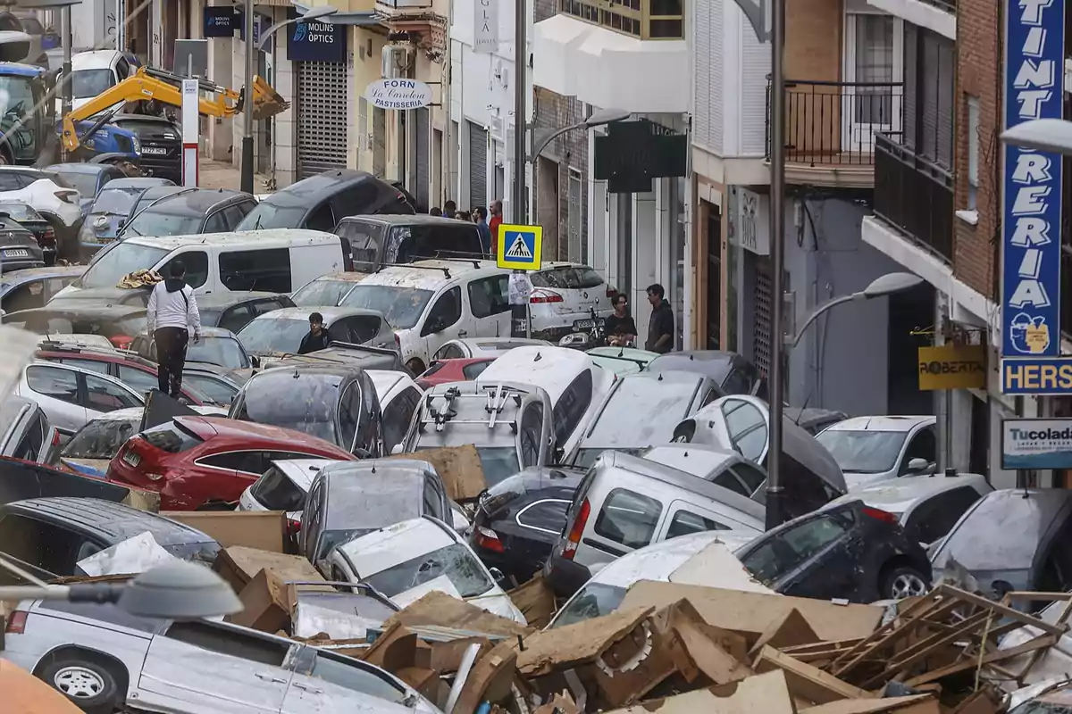 Calle llena de autos amontonados y dañados tras una inundación con personas caminando entre los vehículos y escombros