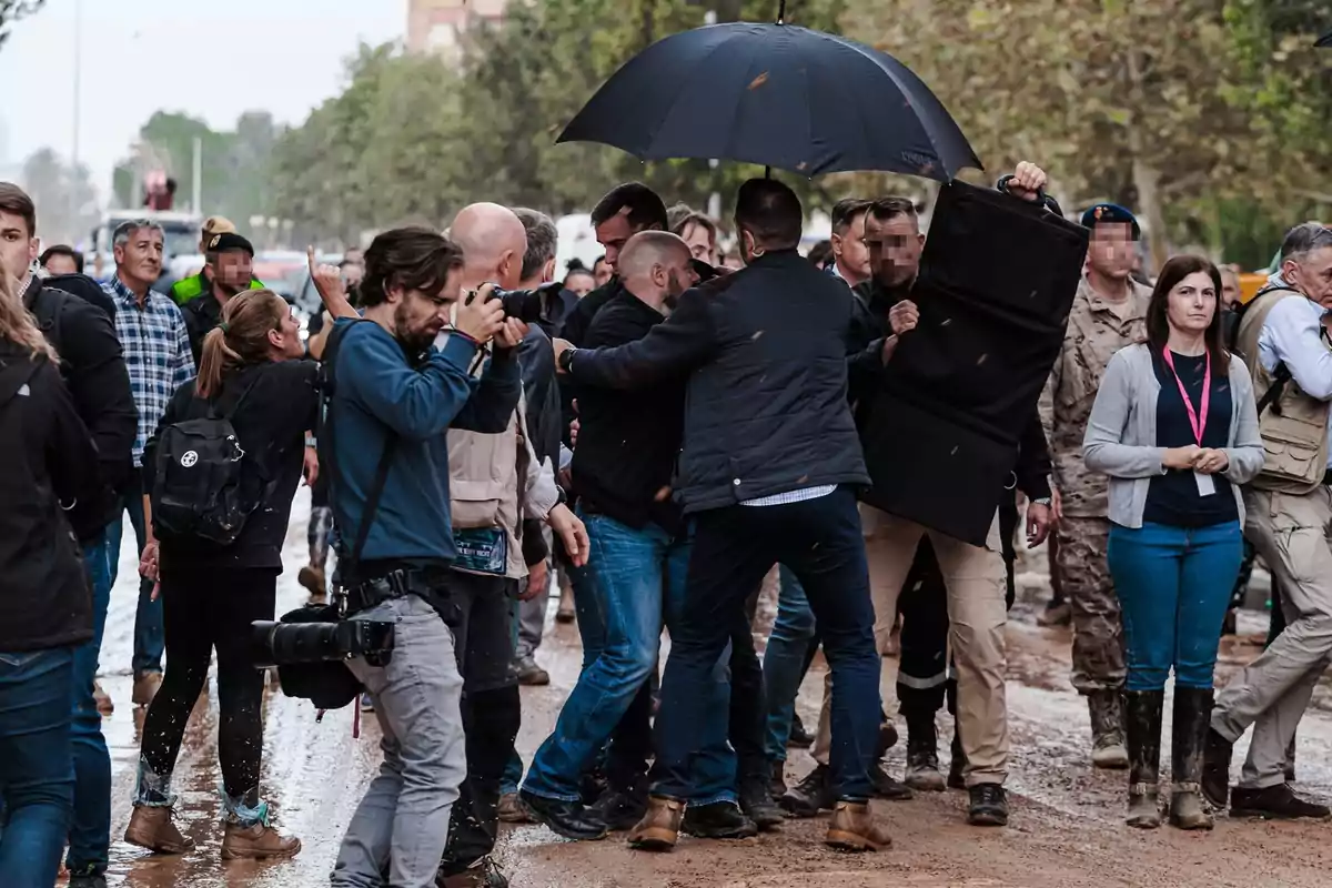 Un grupo de personas, incluyendo fotógrafos, personal de seguridad y militares, se encuentra en una calle embarrada mientras algunos intentan proteger a alguien con un paraguas y un escudo, generando una escena de tensión y movimiento. Un grupo de personas, incluyendo fotógrafos, personal de seguridad y militares, se encuentra en una calle embarrada mientras algunos intentan proteger a alguien con un paraguas y un escudo, generando una escena de tensión y movimiento.