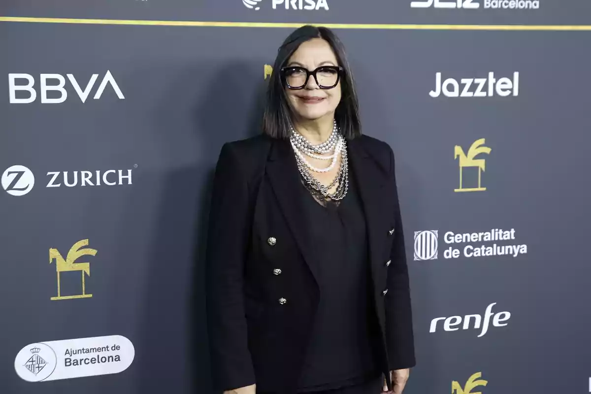 Mujer con gafas grandes y cabello lacio oscuro vestida con un conjunto negro y collares de perlas posando frente a un fondo con logotipos de patrocinadores en un evento