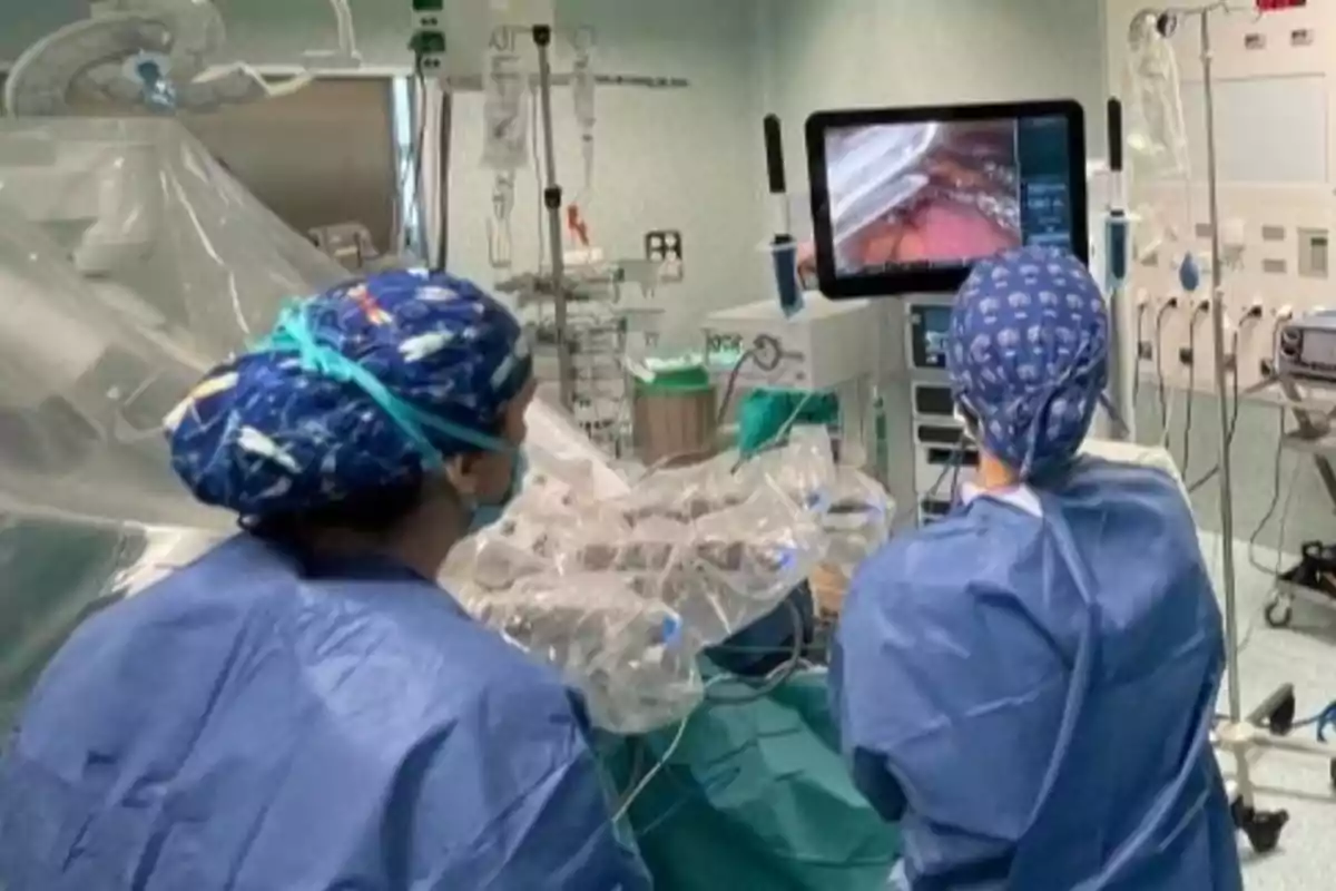 Dos cirujanos con bata y gorro observan una pantalla durante una operación asistida por tecnología en un quirófano moderno Dos cirujanos con bata y gorro observan una pantalla durante una operación asistida por tecnología en un quirófano moderno