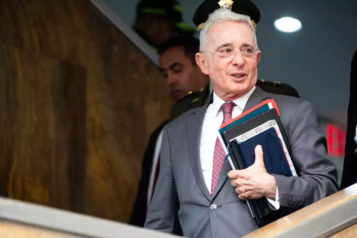 Hombre de cabello canoso con gafas y traje gris sosteniendo varias carpetas y cuadernos mientras baja unas escaleras