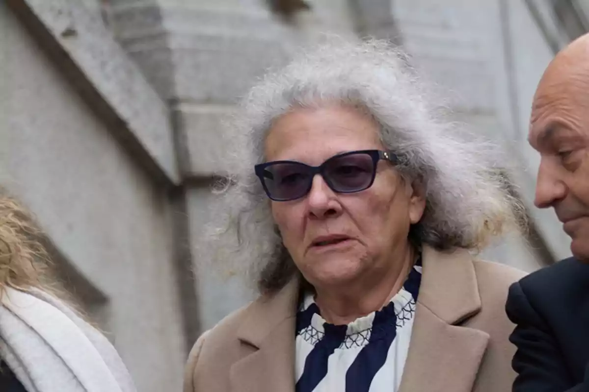 Mujer mayor de cabello canoso y gafas oscuras con expresión seria, vestida con abrigo beige y blusa de rayas, junto a otras personas frente a una pared de piedra Mujer mayor de cabello canoso y gafas oscuras con expresión seria, vestida con abrigo beige y blusa de rayas, junto a otras personas frente a una pared de piedra