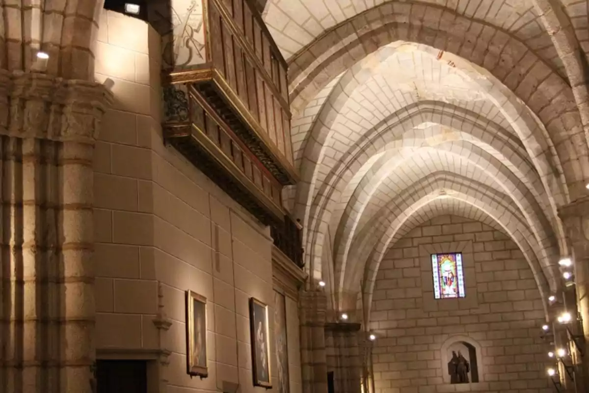 Interior de una iglesia de piedra con techos abovedados, vitrales de colores y cuadros colgados en las paredes