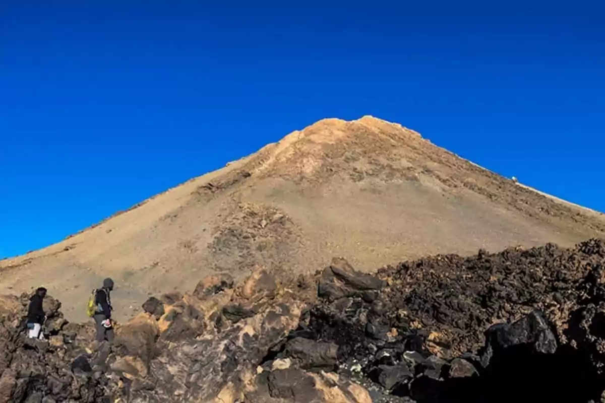 Dos personas caminan entre rocas volcánicas hacia la cima de una montaña bajo un cielo azul despejado
