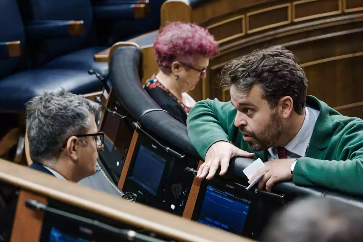 Un hombre con chaqueta verde conversa con otro hombre de gafas en un recinto parlamentario mientras una mujer de cabello corto y teñido está sentada al fondo usando una tableta