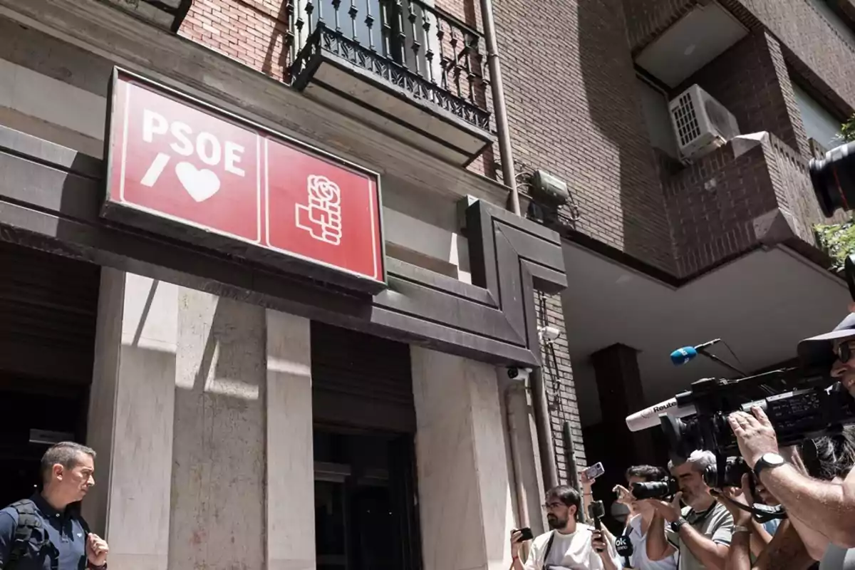 Fachada de un edificio con un cartel rojo del PSOE mientras varios periodistas y camarógrafos graban y toman fotos en la calle