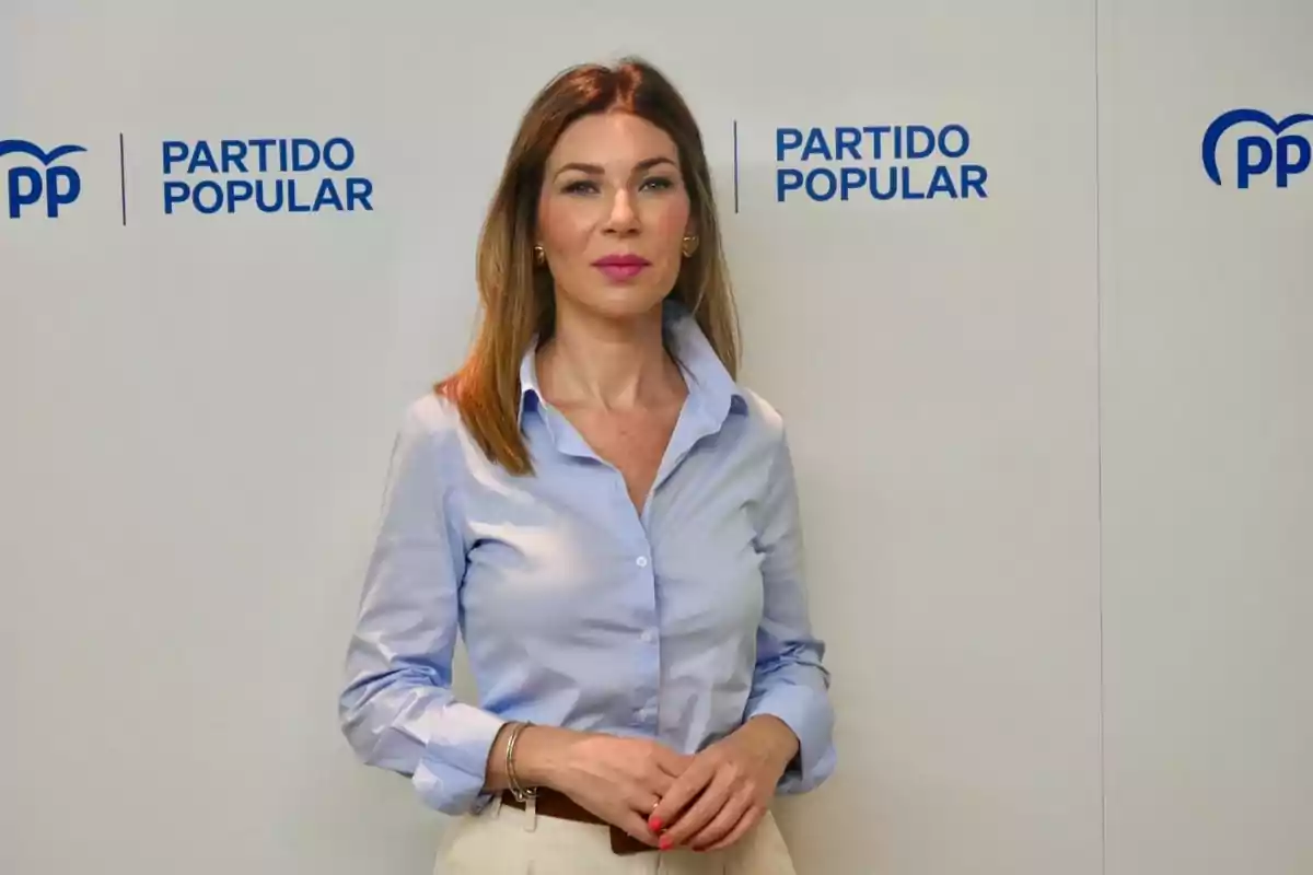 Mujer de cabello castaño y camisa azul claro posando frente a una pared con logotipos del Partido Popular Mujer de cabello castaño y camisa azul claro posando frente a una pared con logotipos del Partido Popular