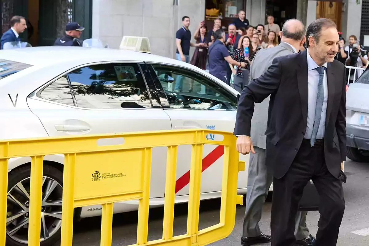 Hombre con traje saliendo de un taxi blanco junto a una valla amarilla mientras varias personas observan y toman fotos al fondo