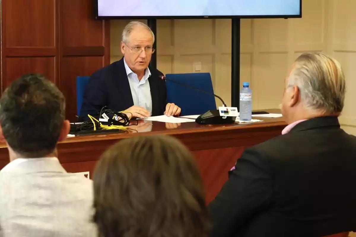 Un hombre mayor con gafas y traje azul habla en una conferencia sentado en una mesa de madera frente a varias personas, con un micrófono y una botella de agua sobre la mesa.