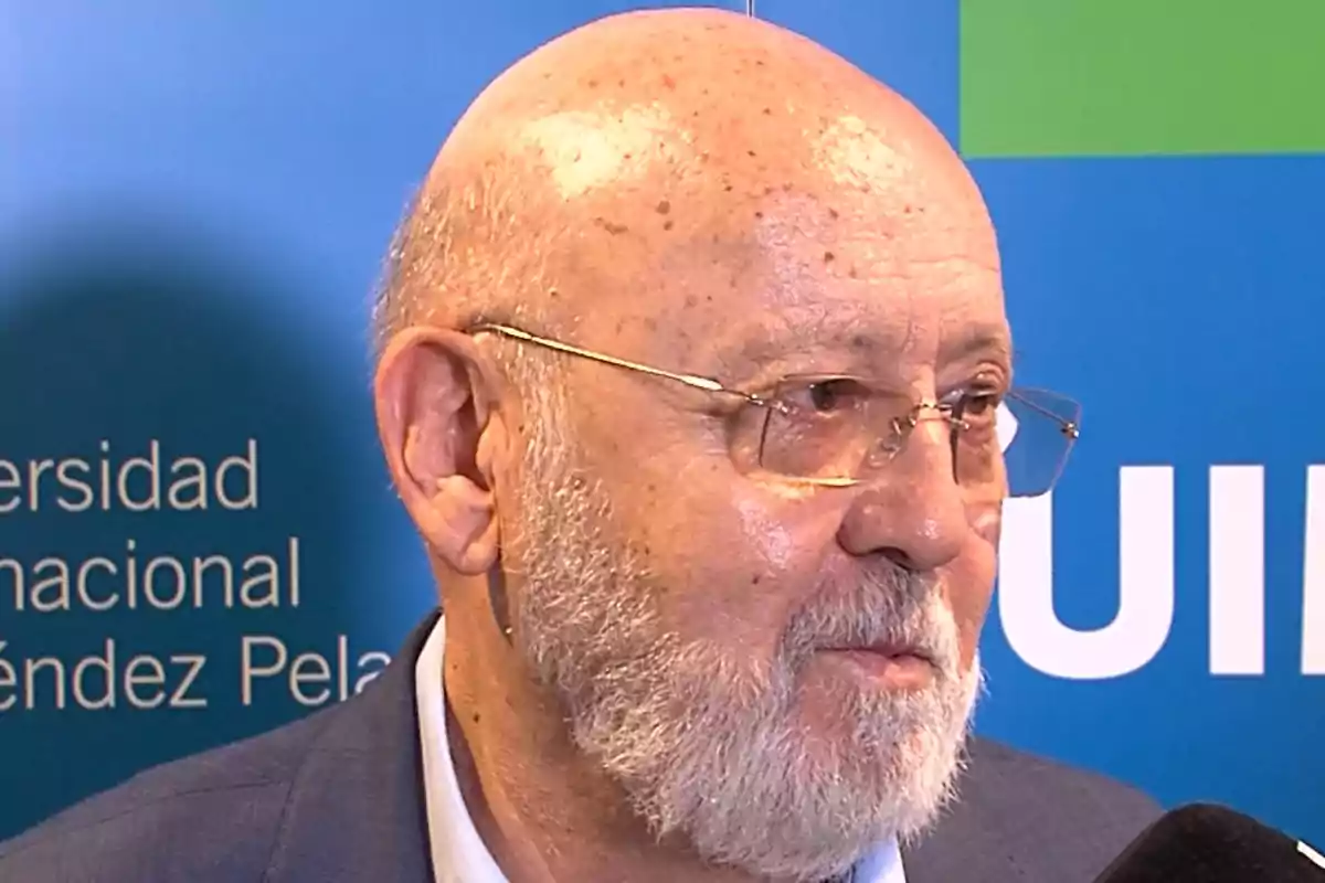 Hombre mayor calvo con barba y gafas siendo entrevistado frente a un fondo azul con texto blanco