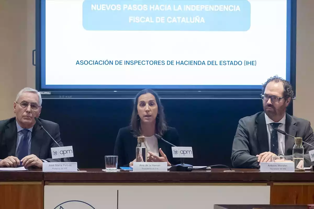 Tres personas con traje sentadas en una mesa durante una conferencia, con un cartel de fondo que menciona la independencia fiscal de Cataluña y la Asociación de Inspectores de Hacienda del Estado