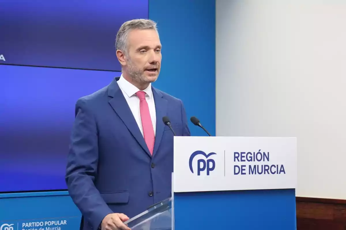 Hombre con traje azul y corbata rosa hablando en un atril con el logo del Partido Popular y el texto Región de Murcia sobre fondo azul Hombre con traje azul y corbata rosa hablando en un atril con el logo del Partido Popular y el texto Región de Murcia sobre fondo azul